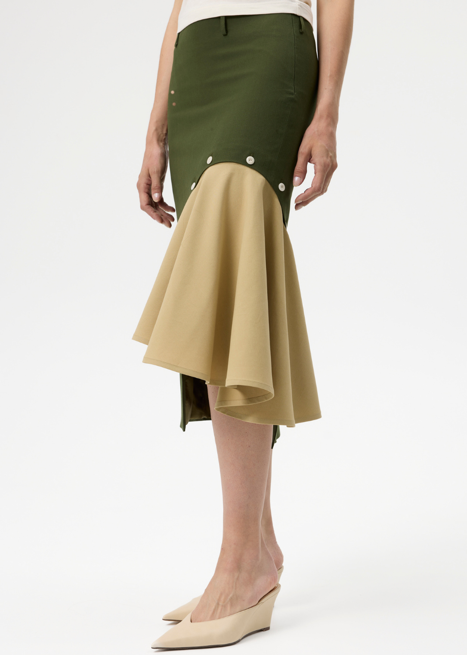 A model wearing an olive green and beige asymmetrical skirt with button details and beige wedge mule heels, photographed against a clean white studio background.