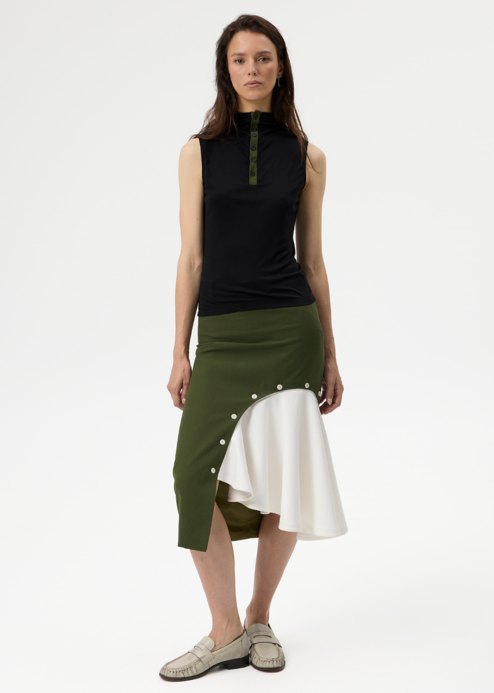 A model stands against a plain white studio background wearing a sleeveless mock-neck top in black with green trim, paired with an asymmetrical midi skirt in olive green and white with decorative buttons, and light-colored loafers.