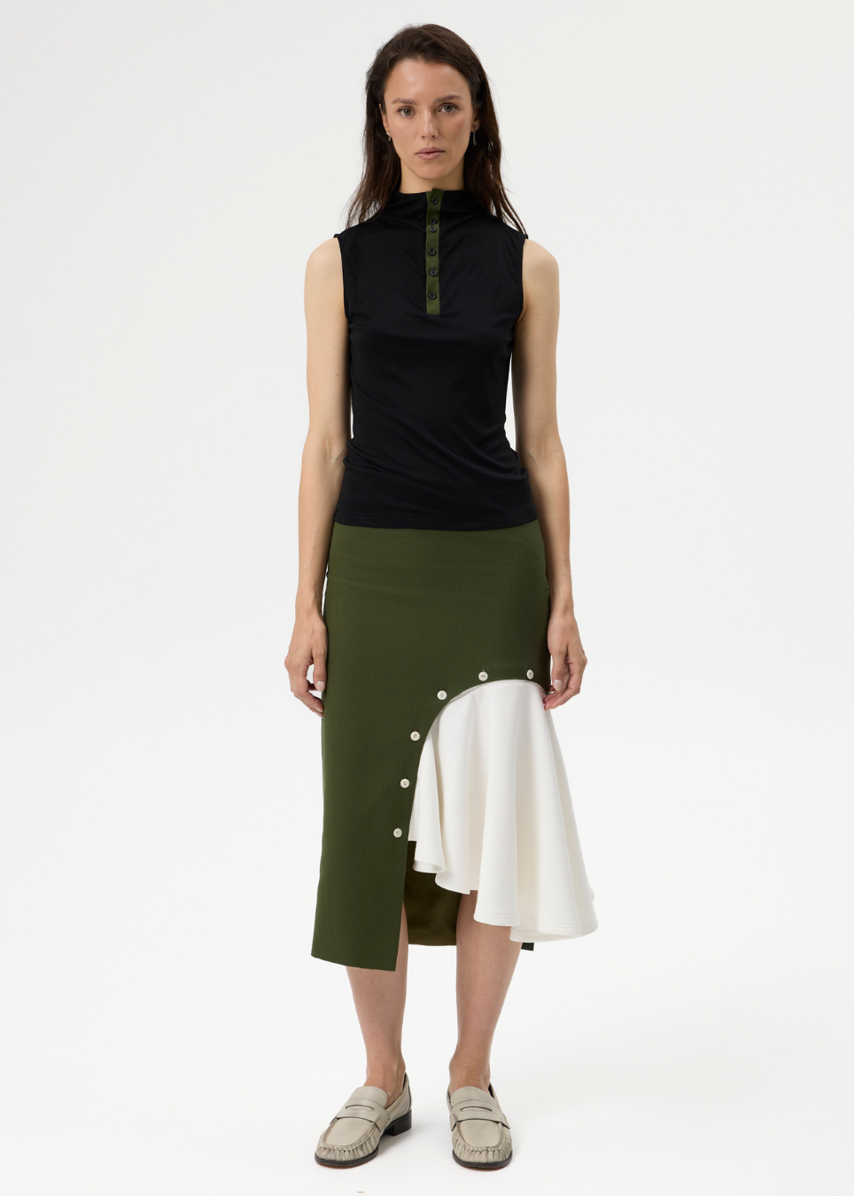A model stands against a plain light gray studio background wearing a sleeveless black mock-neck top with green buttons and an asymmetrical olive green skirt with a white ruffled overlay and white buttons.