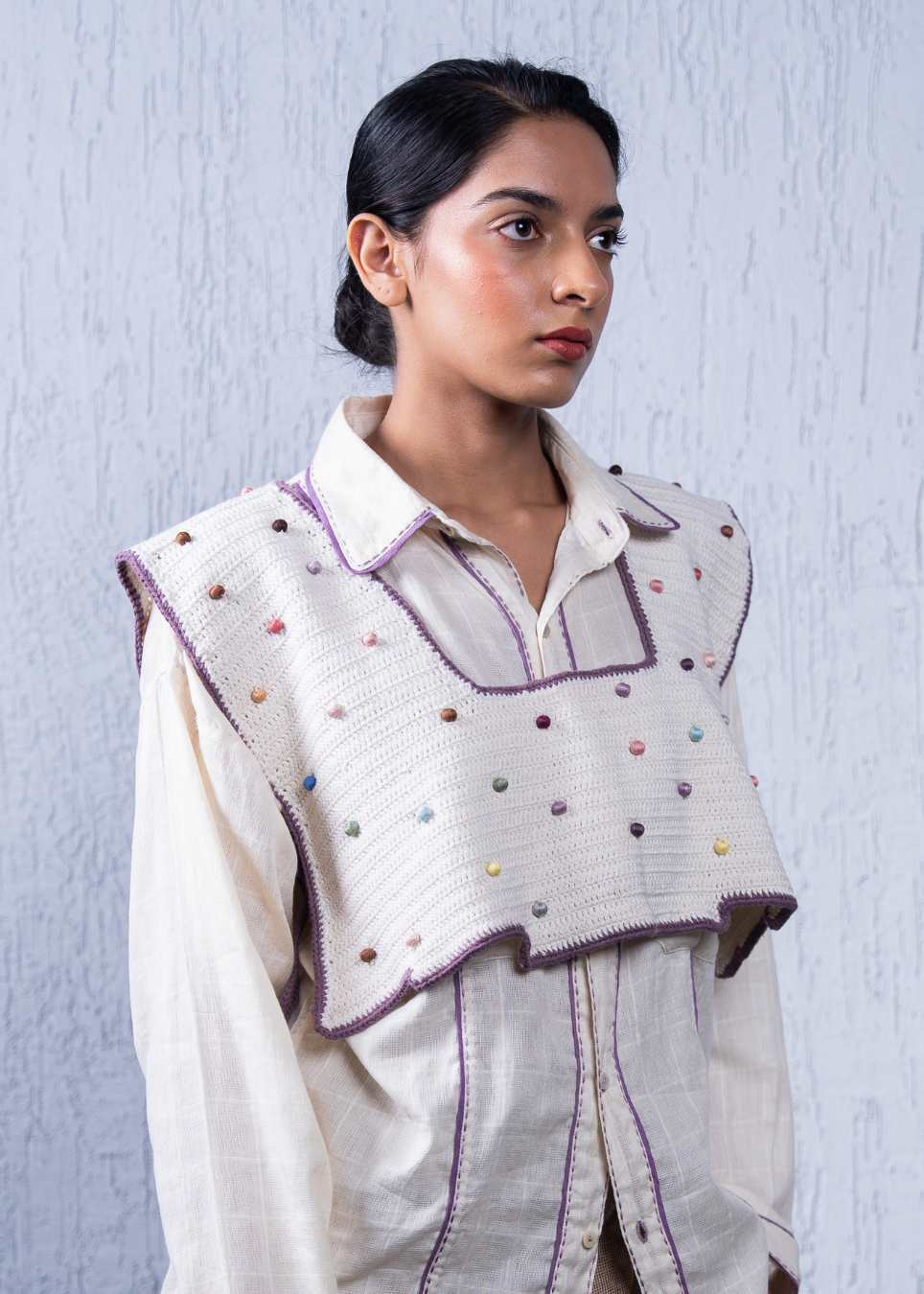 A woman wearing a cream-colored embroidered vest over a matching cream shirt, set against a textured light blue-gray background.
