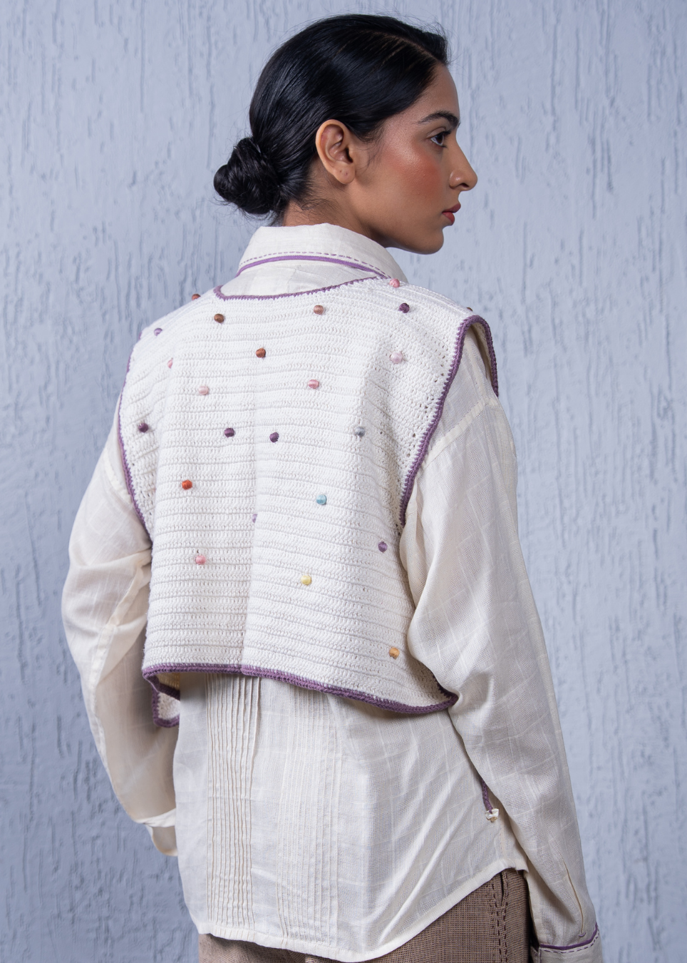 A woman shown from the back wearing a cream-colored textured vest with purple trim and small multicolored pom-pom embellishments, layered over a cream shirt.