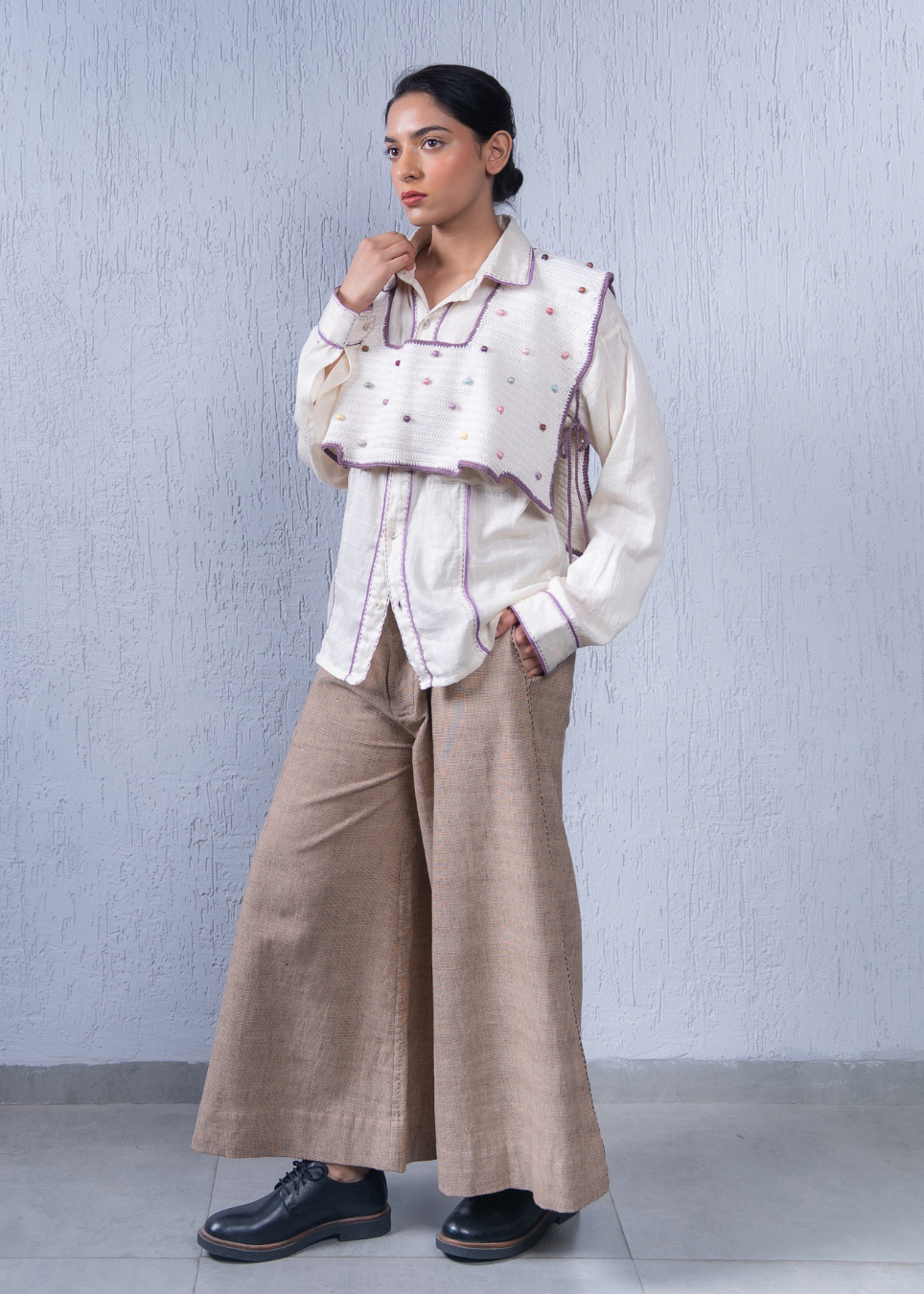 A woman models a white long-sleeve button-up shirt with purple trim and a matching white cropped vest decorated with small purple and pink embroidered dots, paired with wide-leg beige trousers and black shoes, set against a textured light gray wall.