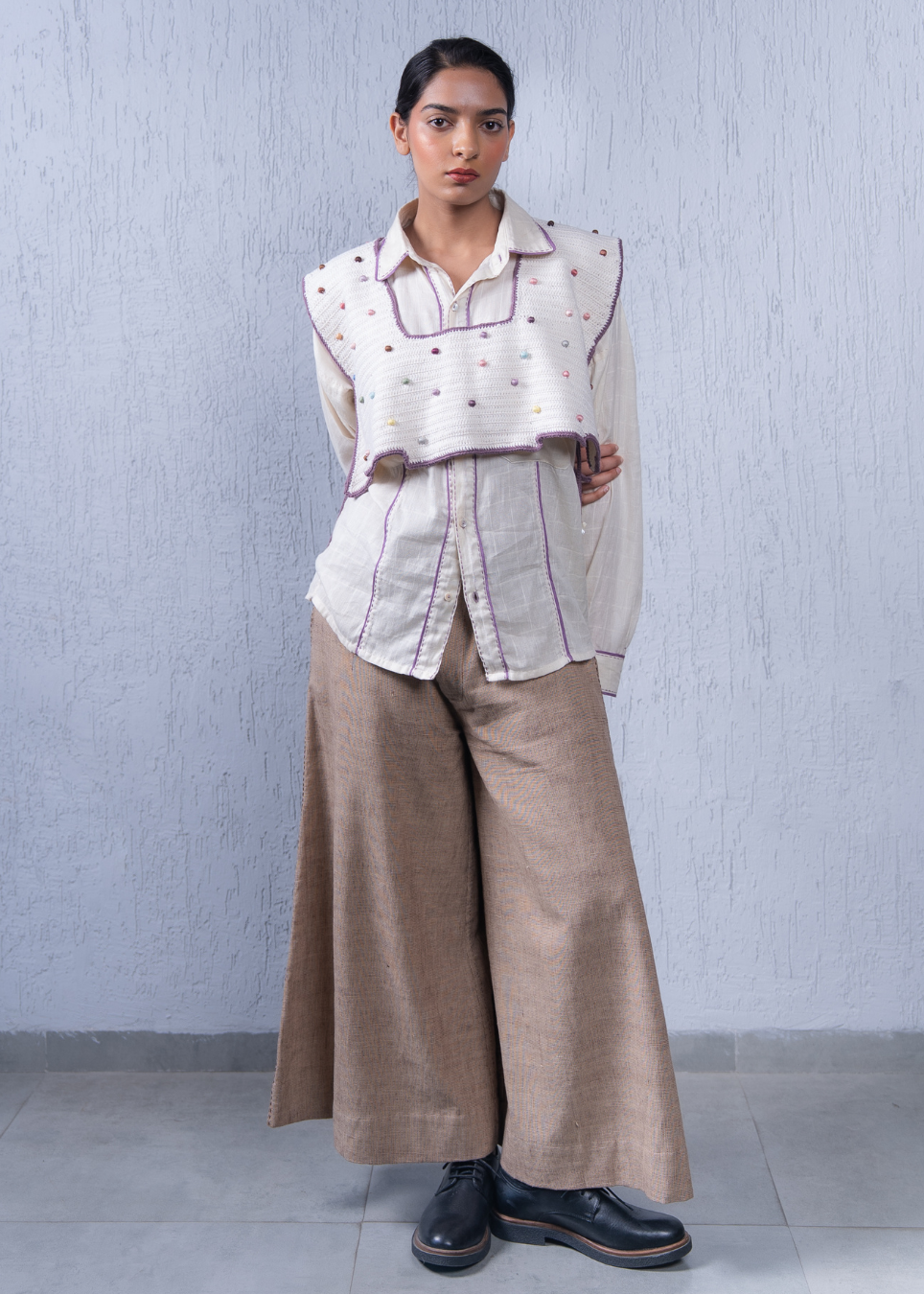 A model stands against a textured light gray wall wearing a cream-colored sleeveless embroidered vest over a matching button-up shirt, paired with wide-leg beige trousers and black leather shoes.