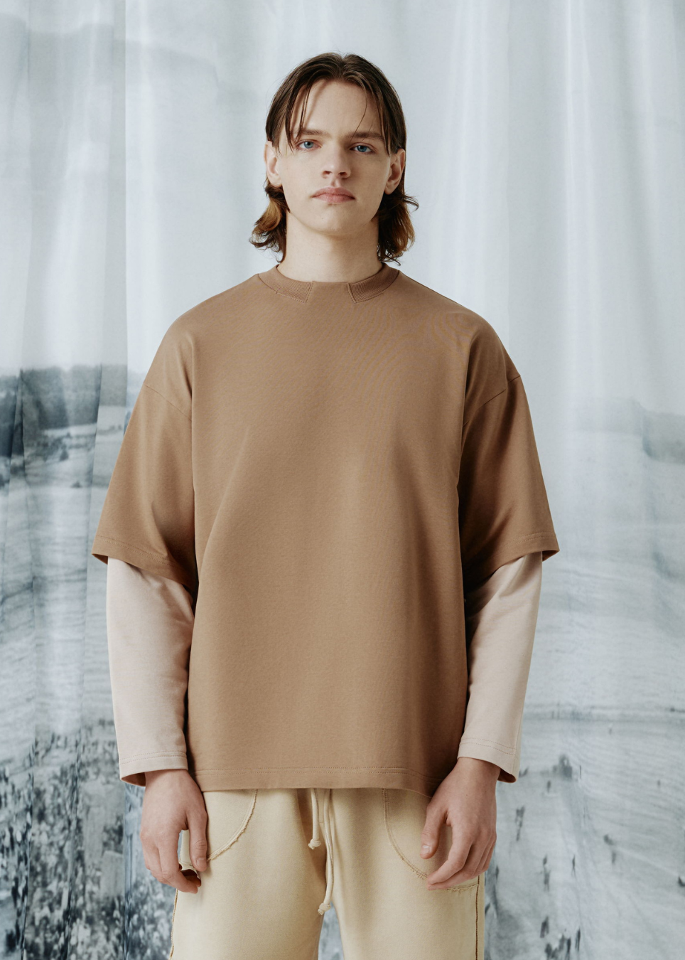 A model wearing a beige short-sleeve mock neck t-shirt with integrated cream-colored long sleeves, paired with beige drawstring pants, standing against a soft, misty landscape backdrop.