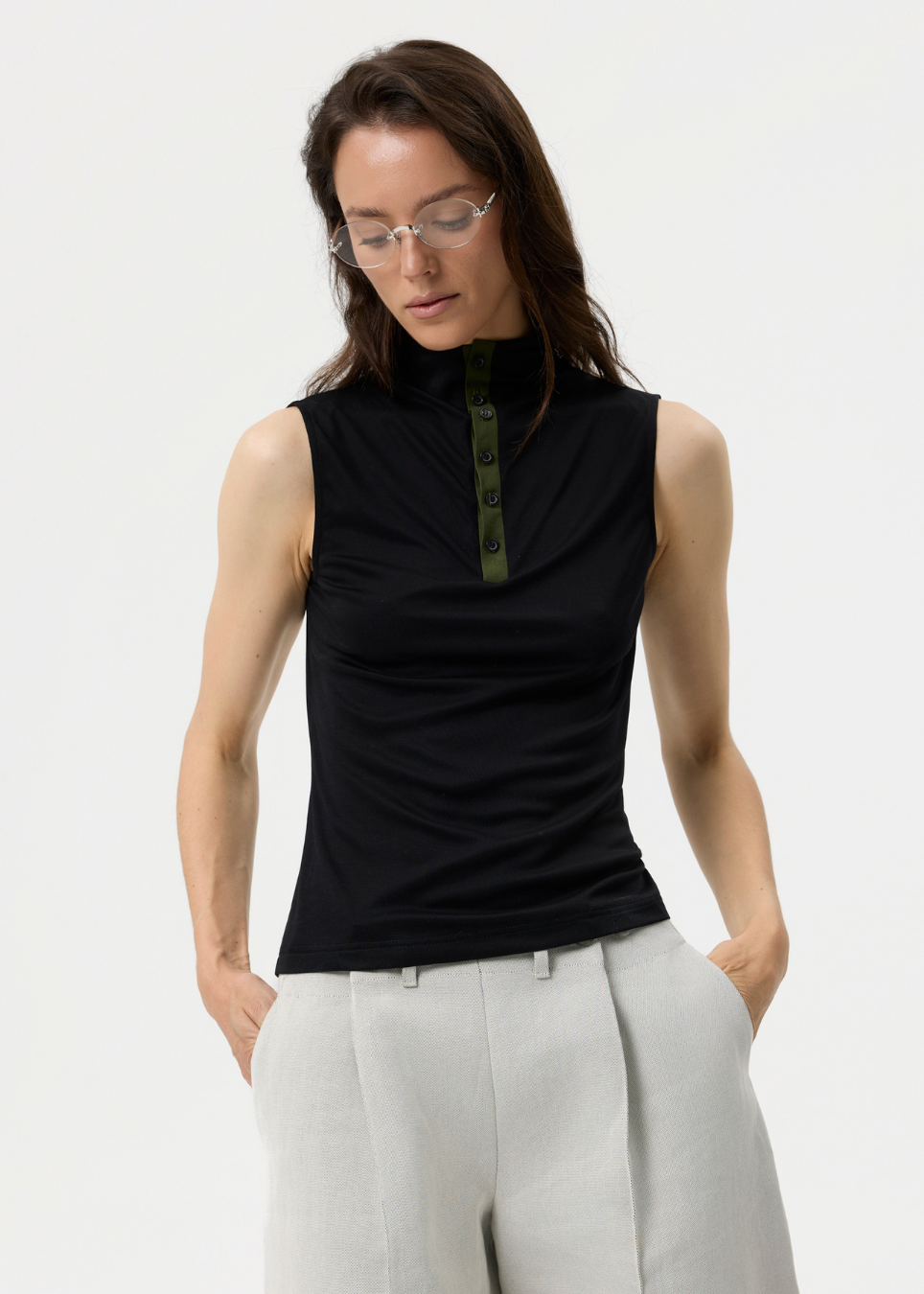 A woman wearing a black sleeveless mock-neck top with a green button placket and light gray trousers, photographed against a clean white studio background.