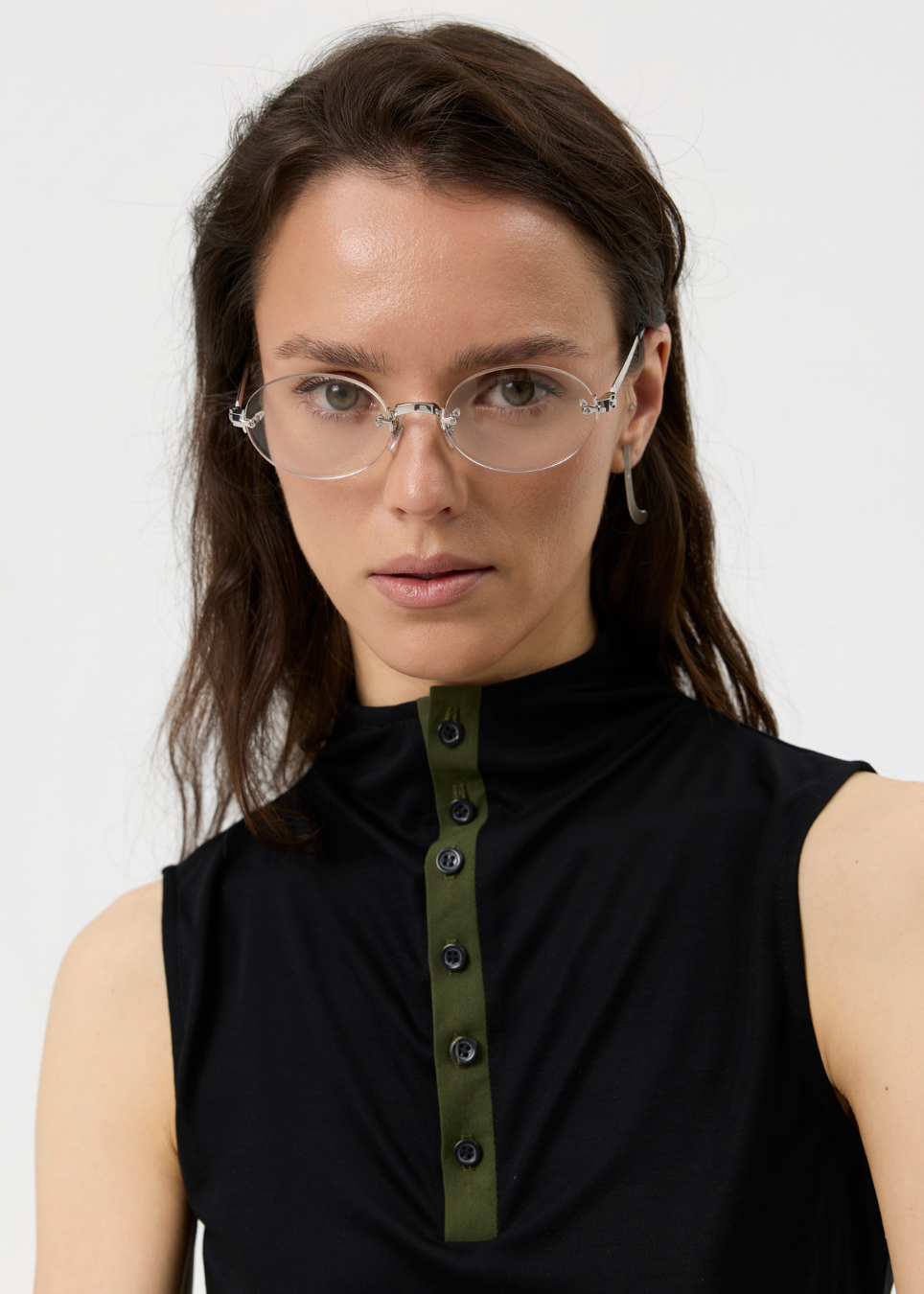 A close-up of a woman wearing clear-framed eyeglasses and a sleeveless black top with an olive green button placket, against a plain light background.
