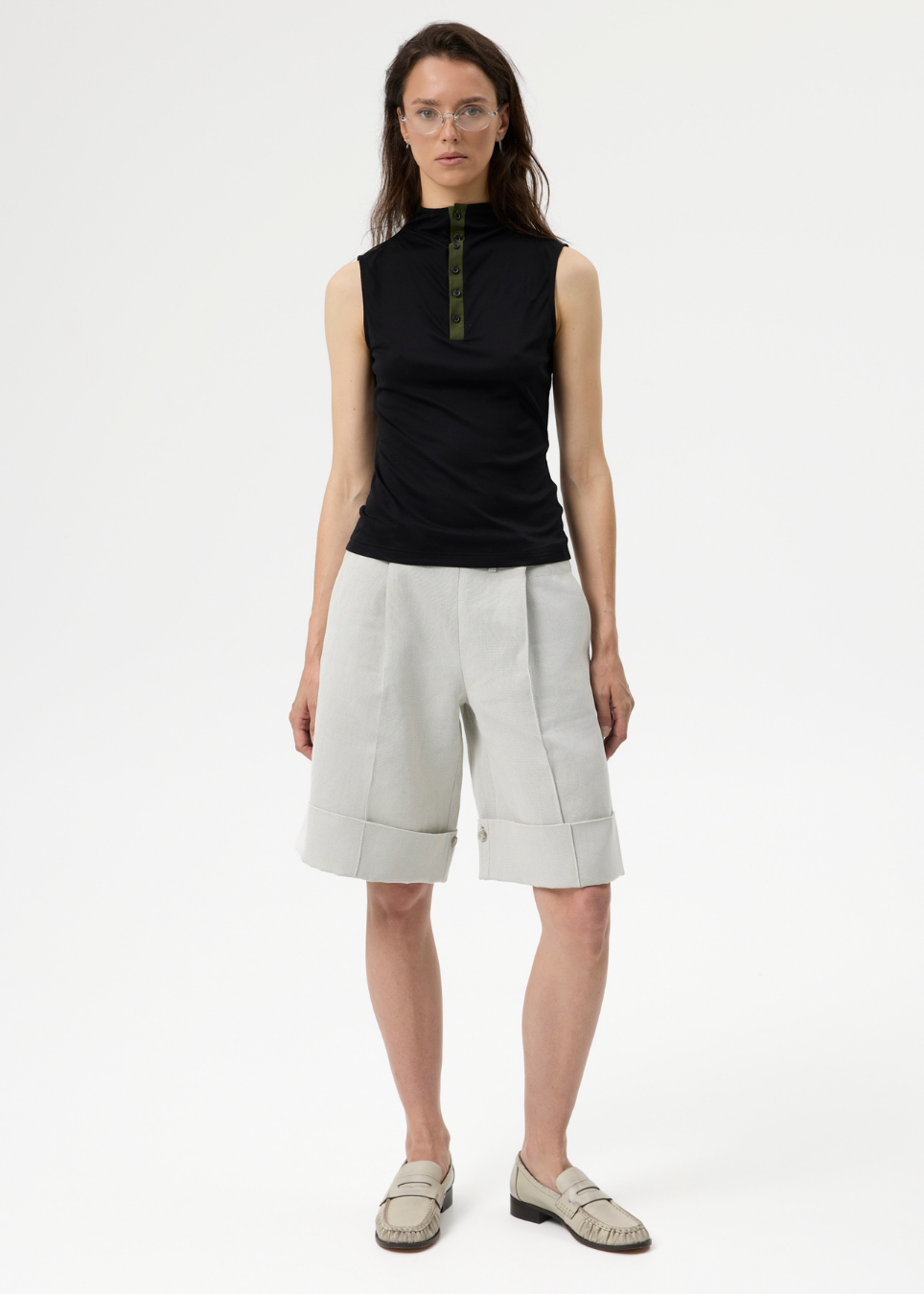 A model stands against a plain white studio background wearing a sleeveless black top with a green zipper detail, light beige pleated shorts, and beige loafers.