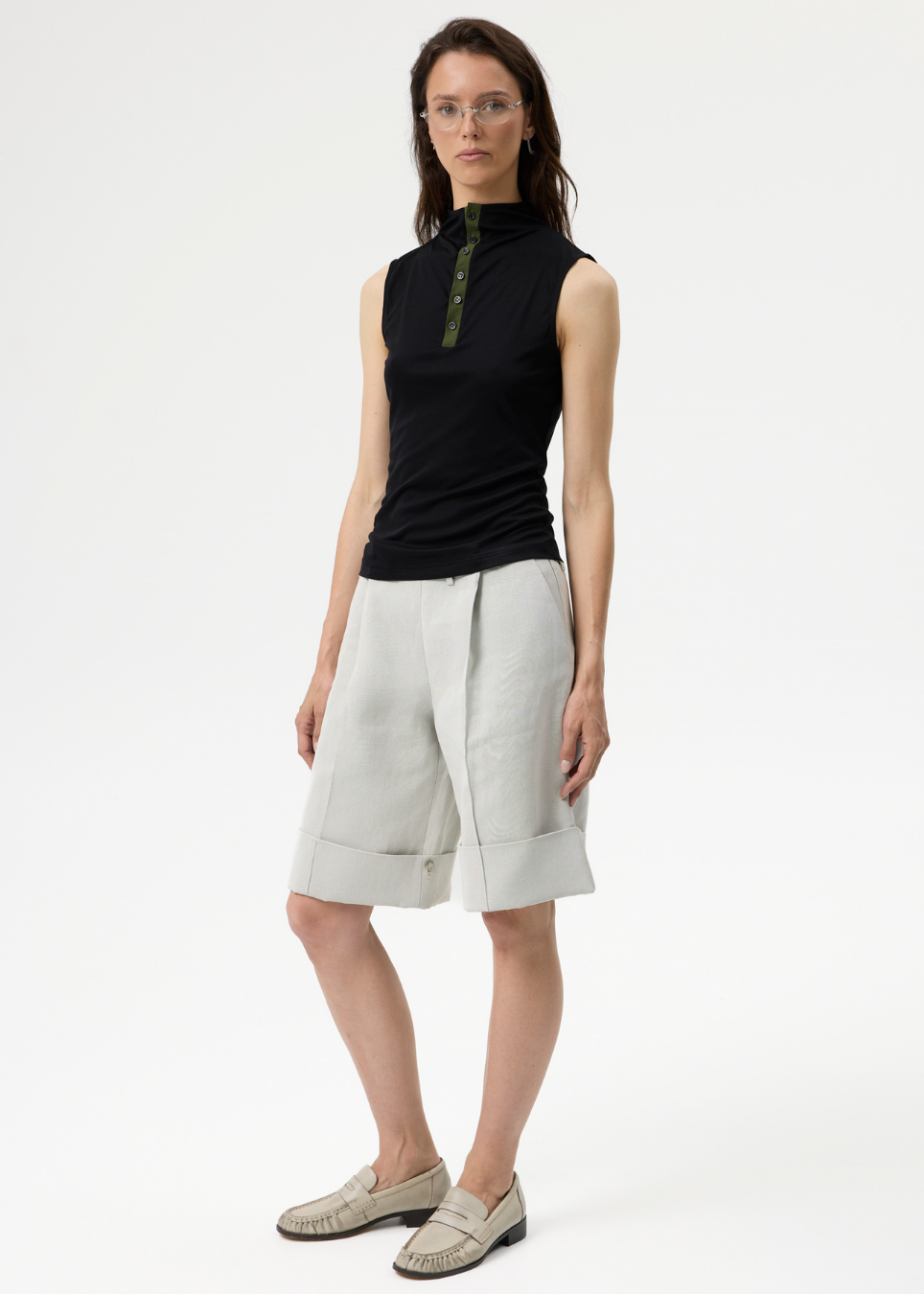 A model stands against a plain white studio background wearing a sleeveless black top with a green zipper detail at the collar, light beige pleated shorts, and beige loafers.
