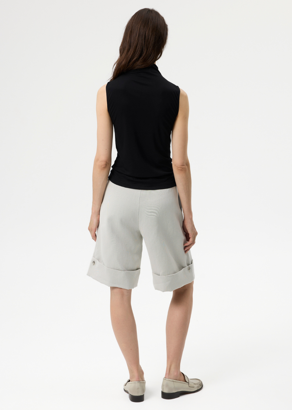 A woman shown from the back wearing a black sleeveless mock-neck top and light gray wide-leg shorts, paired with gray slip-on shoes, against a plain white studio background.
