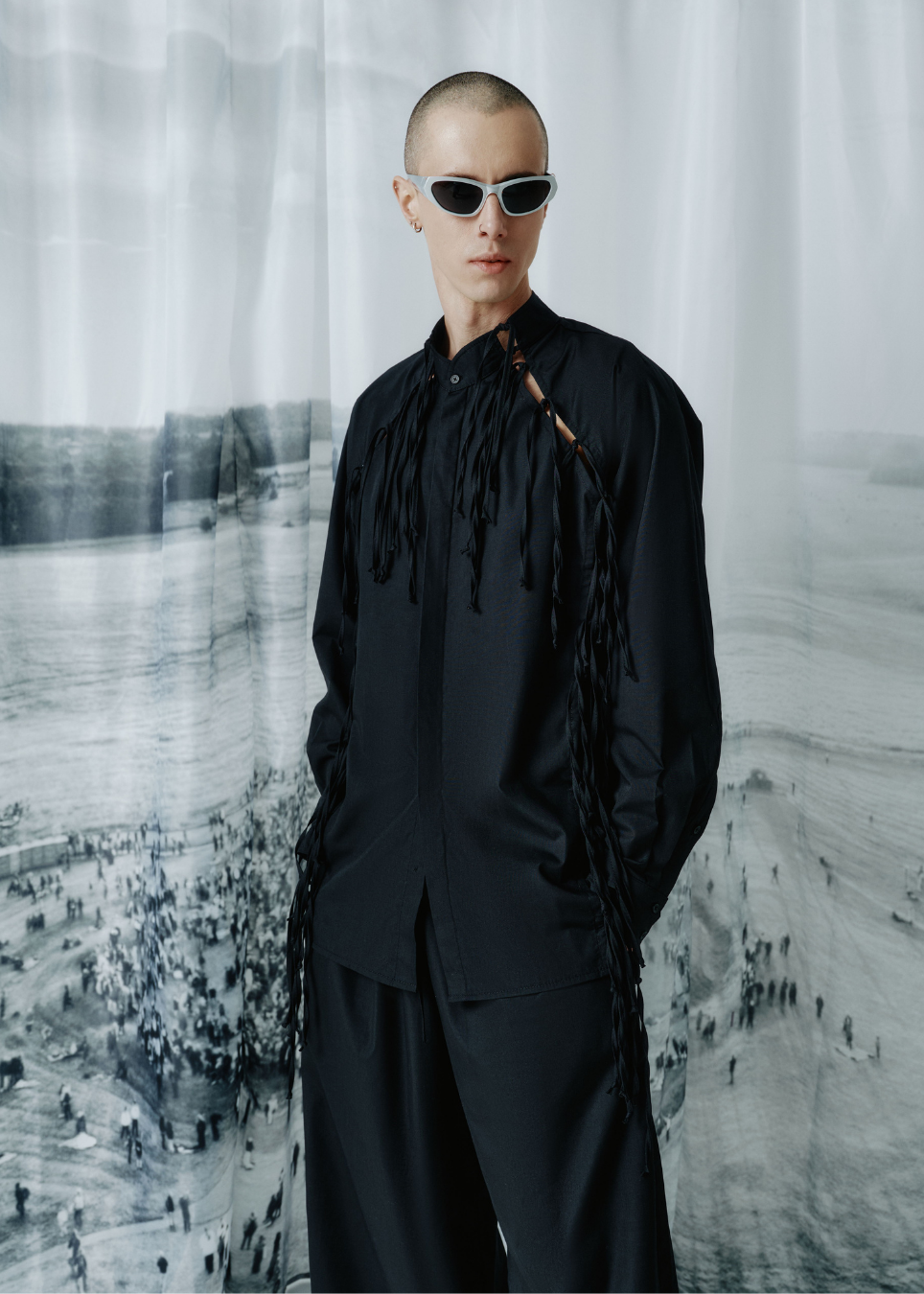 A model with a shaved head wearing a black long-sleeve fringed shirt and matching wide-leg trousers, styled with white-framed sunglasses, standing in front of a large black-and-white beach photograph.