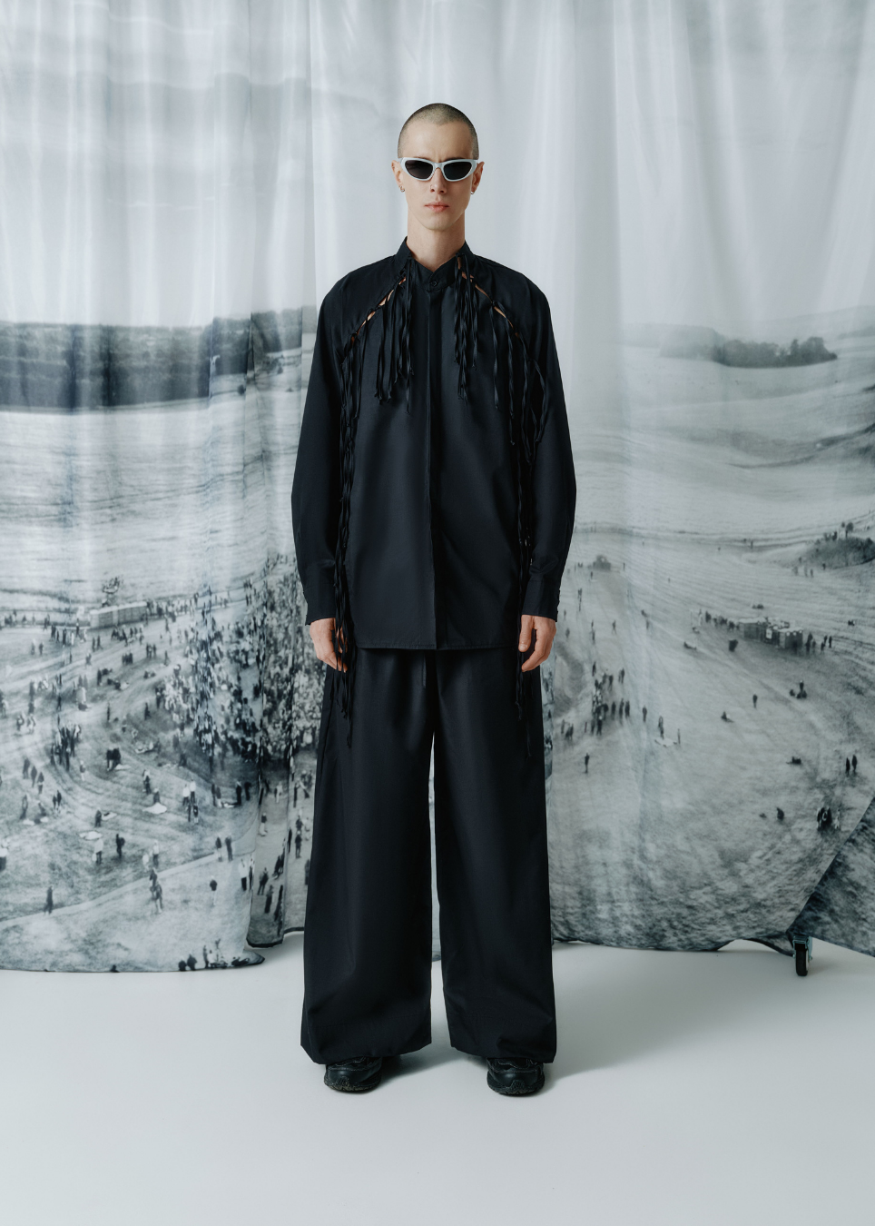 A model stands facing forward wearing a black long-sleeve shirt with dramatic fringe details down the front and matching wide-leg black pants, set against a large black-and-white landscape backdrop.