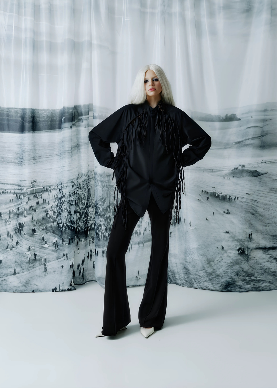 A model with platinum blonde hair stands in a studio wearing a black long-sleeve shirt with dramatic fringe and black flared pants. The background is a large black-and-white scenic photograph printed on a draped fabric backdrop.