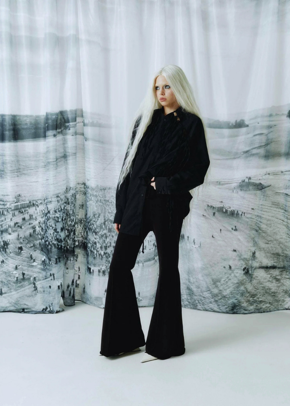 A model with long platinum blonde hair stands in a studio wearing a black shirt and matching black flared pants. The background features a large, draped black and white landscape photograph.