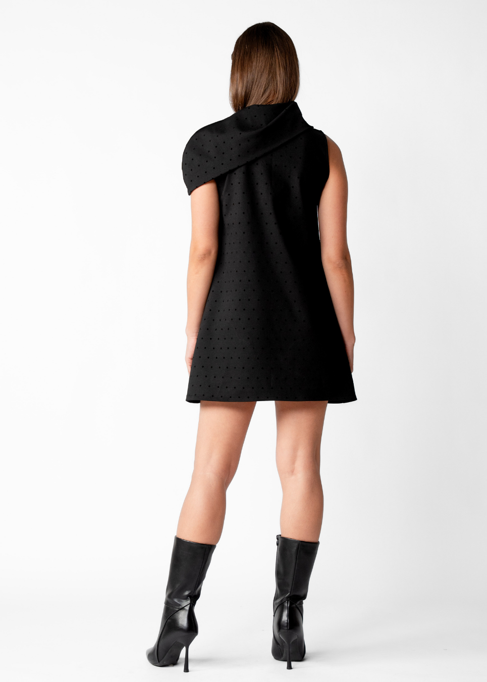 A woman shown from the back wearing a black sleeveless dress with a draped, asymmetrical collar and a subtle textured pattern, paired with black heeled boots against a plain white studio background.