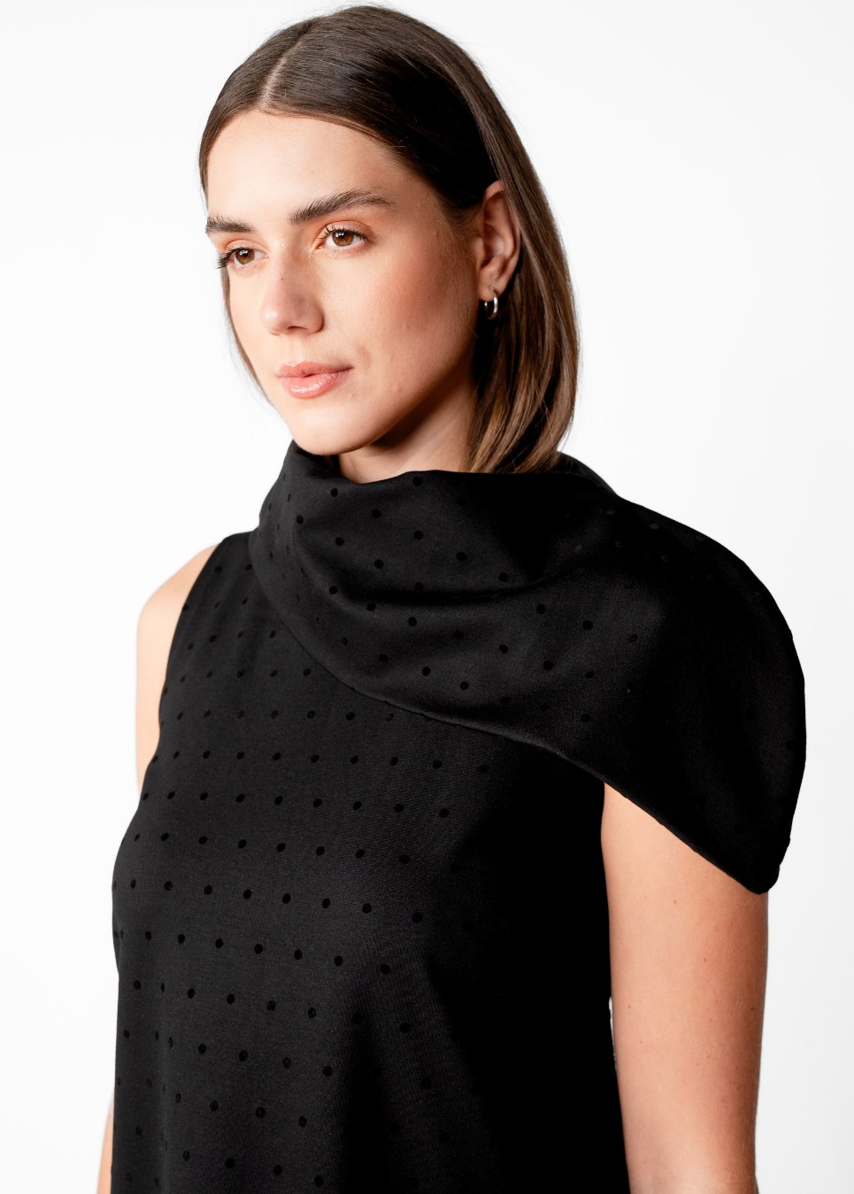 A woman wearing a black sleeveless top with a high draped mock neck and a subtle perforated dot texture, photographed against a plain white studio background.