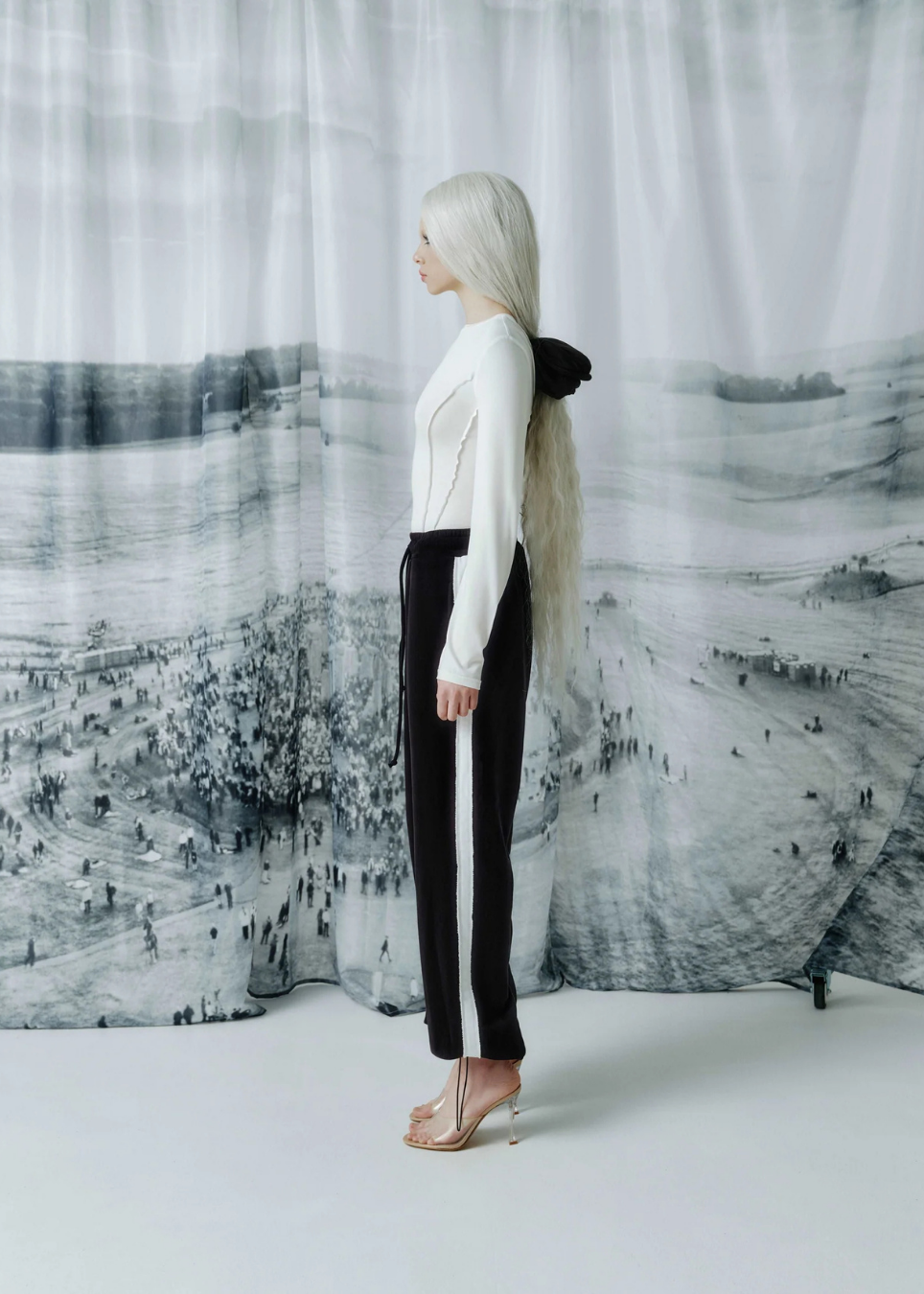 A model with long platinum blonde hair stands in profile wearing a white ribbed long-sleeve top and black wide-leg trousers with a white side stripe, posed in front of a large black and white landscape backdrop.