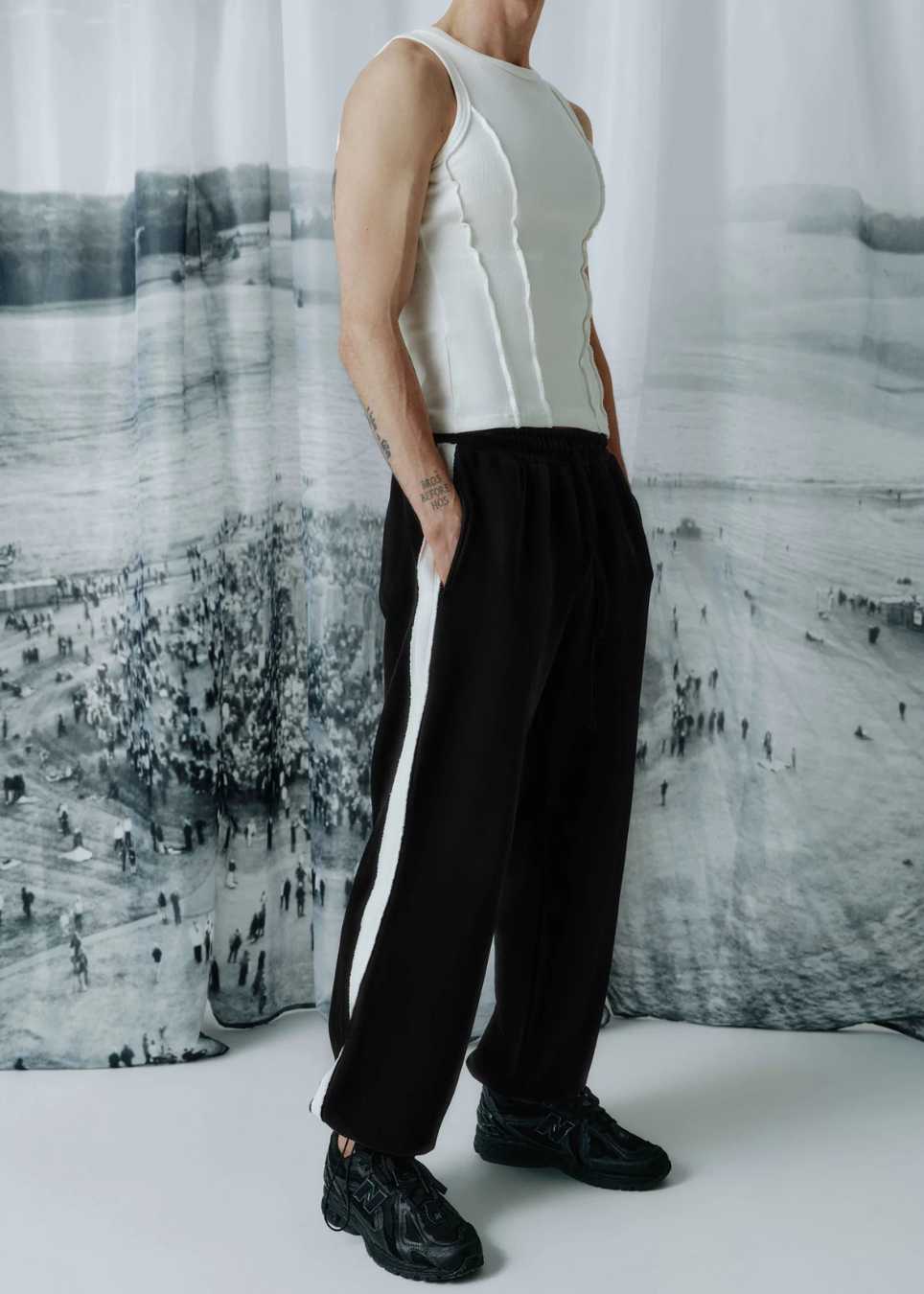 A model wearing a sleeveless white top and black wide-leg track pants with white side stripes, standing in front of a black and white scenic backdrop.