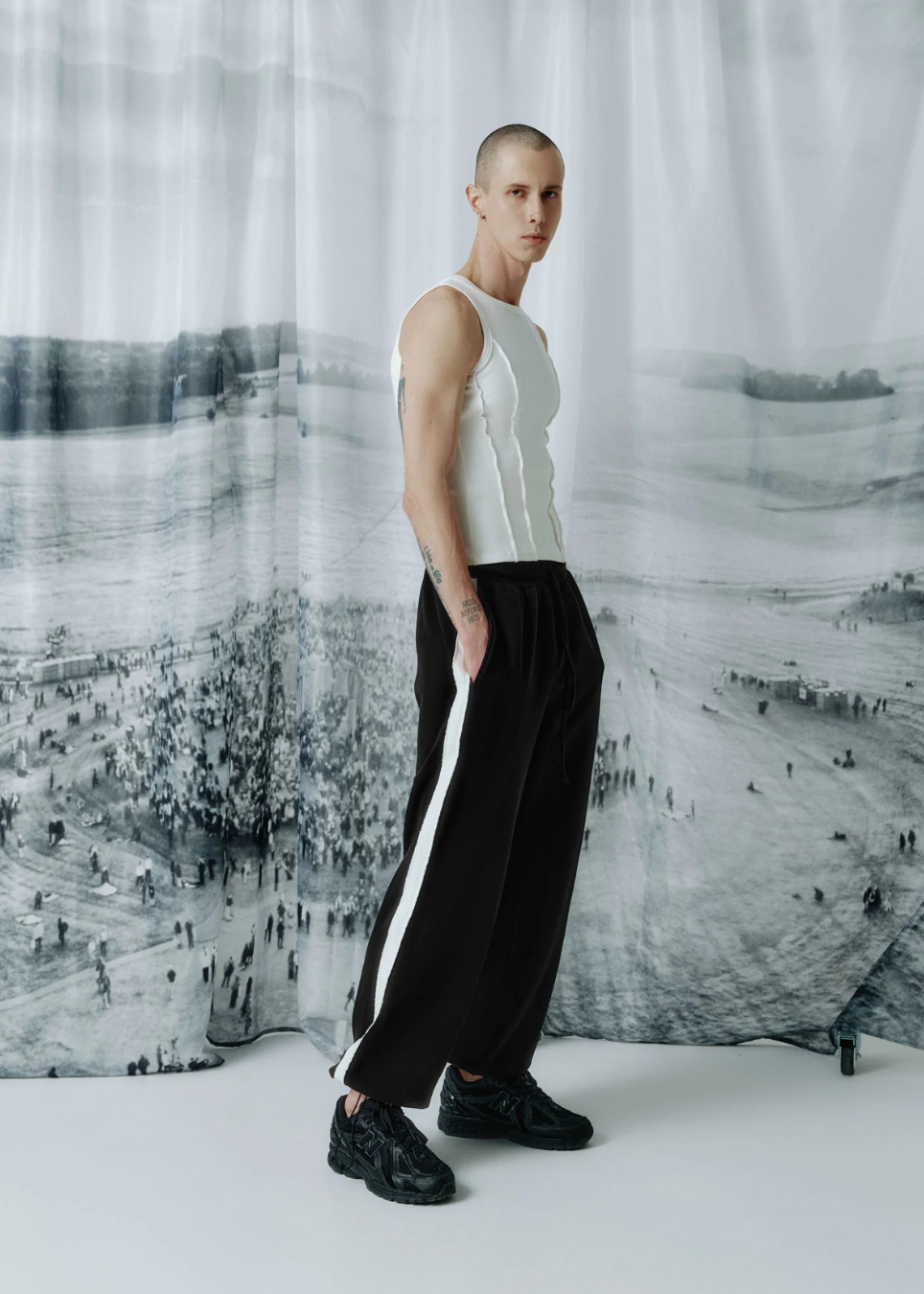 A model stands in profile wearing a sleeveless white top and loose black pants with a bold white side stripe, posed against a large black-and-white scenic backdrop.
