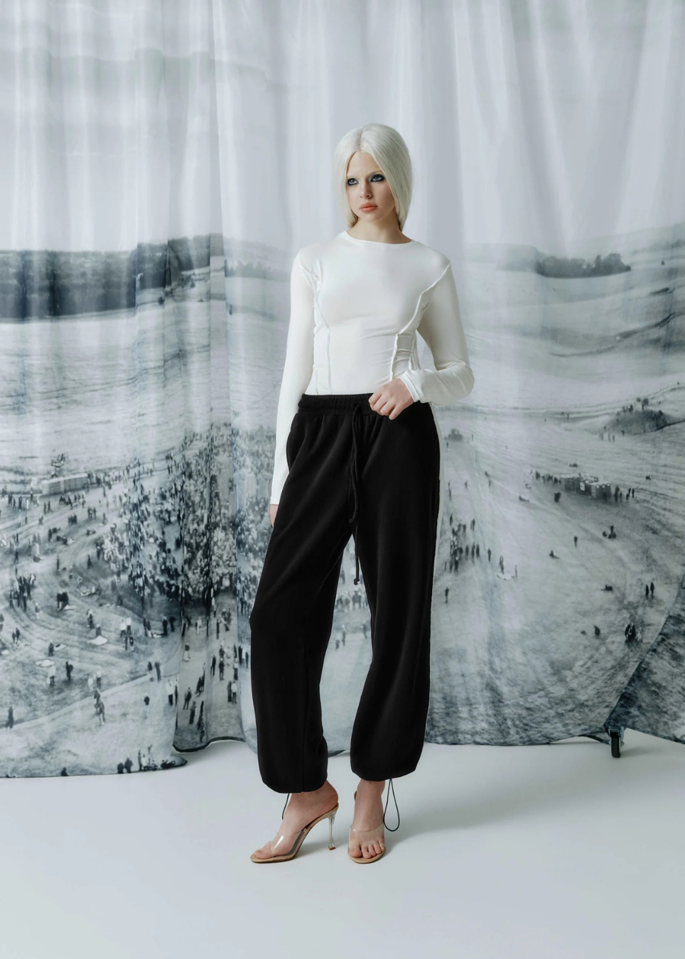 A model with platinum blonde hair stands in front of a black and white landscape print curtain, wearing a white long-sleeve mock-neck top with ruffle details, black wide-leg cropped pants, and clear high-heeled sandals.