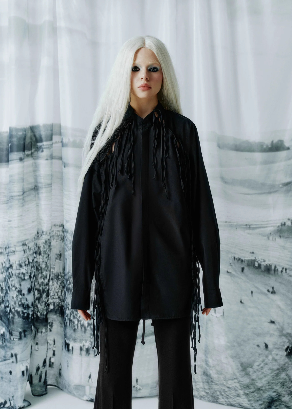 A model with long platinum blonde hair stands wearing a black long-sleeve shirt with dramatic fringe details and black pants, posed in front of a large black and white landscape photograph backdrop.