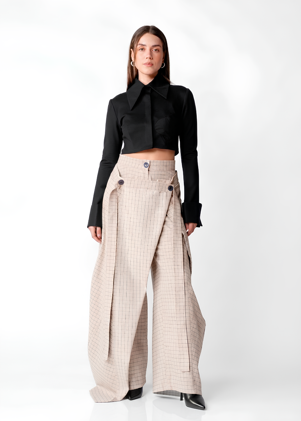 A model stands against a white studio background wearing a black long-sleeve cropped shirt with a high collar and wide-leg beige plaid trousers.