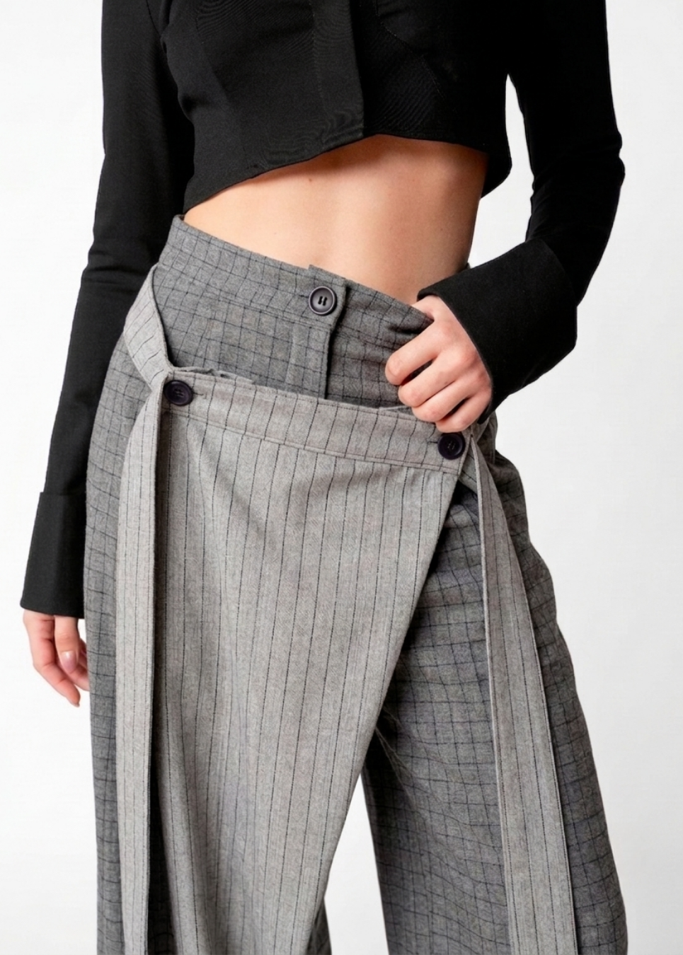 Close-up of a model wearing olive pinstriped wide-leg trousers with a unique buttoned waist and long tie details, paired with a black cropped long-sleeve top against a white studio background.