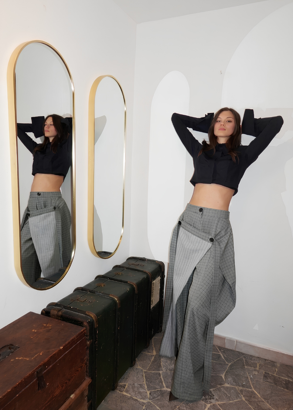 A woman poses in a room wearing a black long-sleeve crop top and a high-waisted, wide-leg olive plaid pant-skirt. She stands next to two tall arched mirrors with gold frames, reflecting her figure, with vintage dark green suitcases on the floor.
