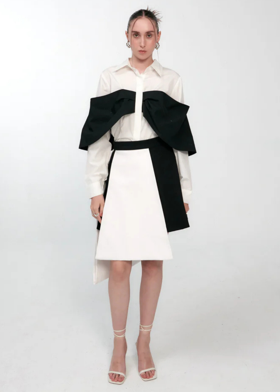 A model wearing a black and white asymmetrical dress with a large bow detail on the shoulders, a white collared shirt underneath, and clear high-heeled sandals, photographed against a light gray studio background.