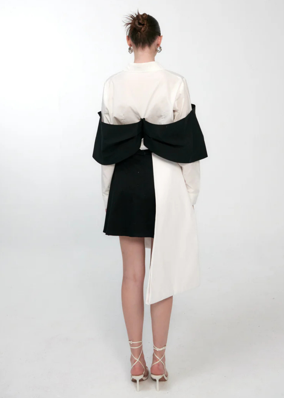 A model shown from the back wearing a unique asymmetrical black and white dress with a large bow detail across the shoulders, paired with white lace-up high heels, against a plain white studio background.