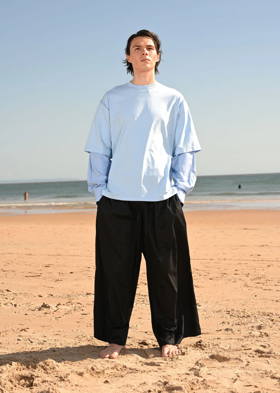 A man standing on a sandy beach wearing an oversized light blue long-sleeve shirt and loose black pants, with the ocean and blue sky in the background.