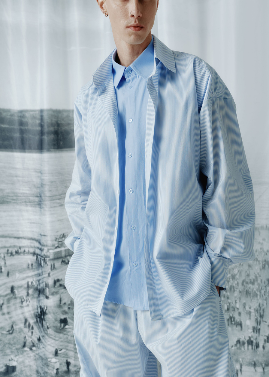 A model wearing a light blue button-down shirt and matching light blue trousers, standing in front of a large black-and-white scenic print.