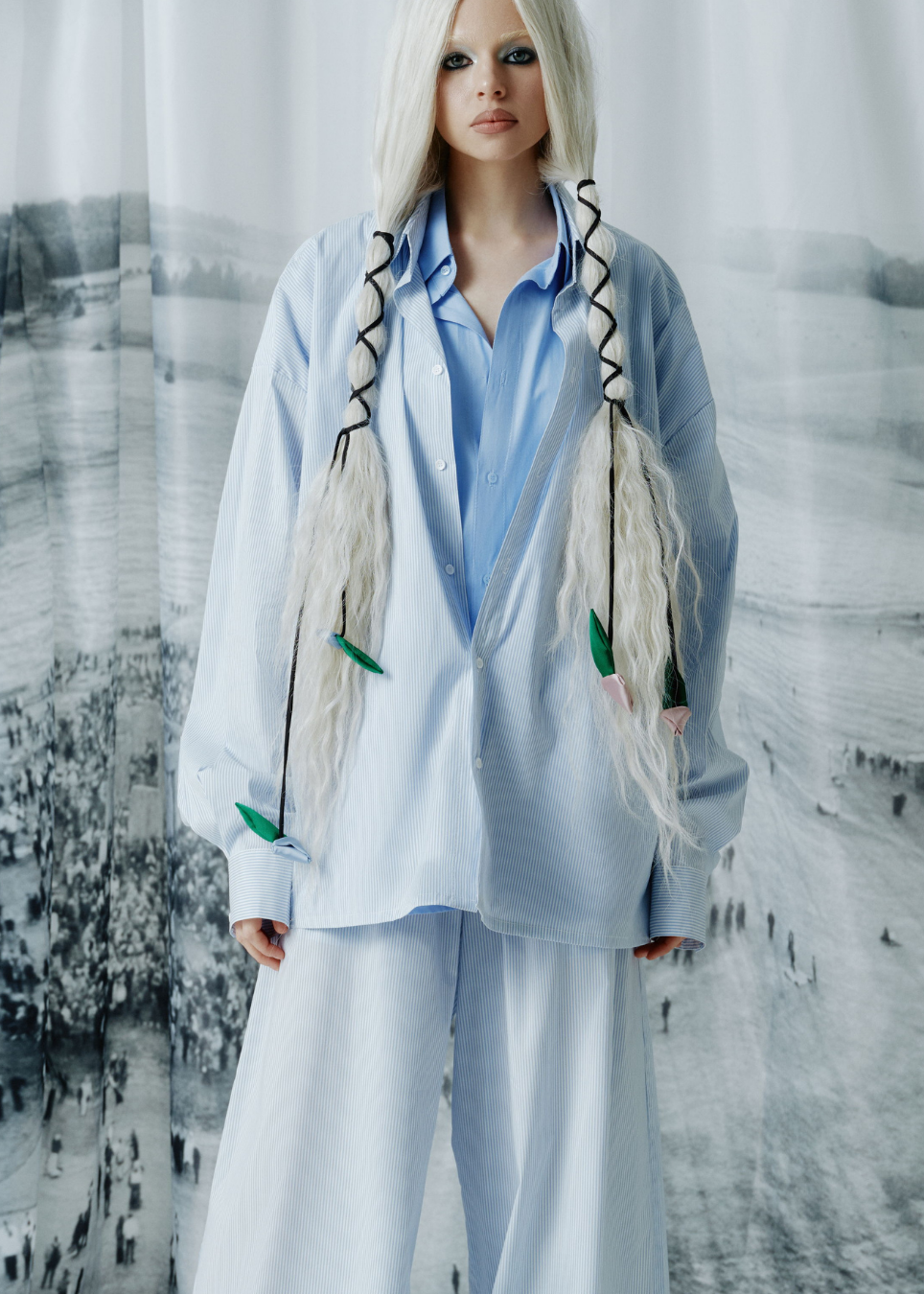 A model with long platinum blonde hair styled in two braids wearing a light blue button-up shirt, matching wide-leg trousers, and a light blue oversized overshirt decorated with green leaf-like accents and black braided details, posed in front of a large black and white landscape tapestry.
