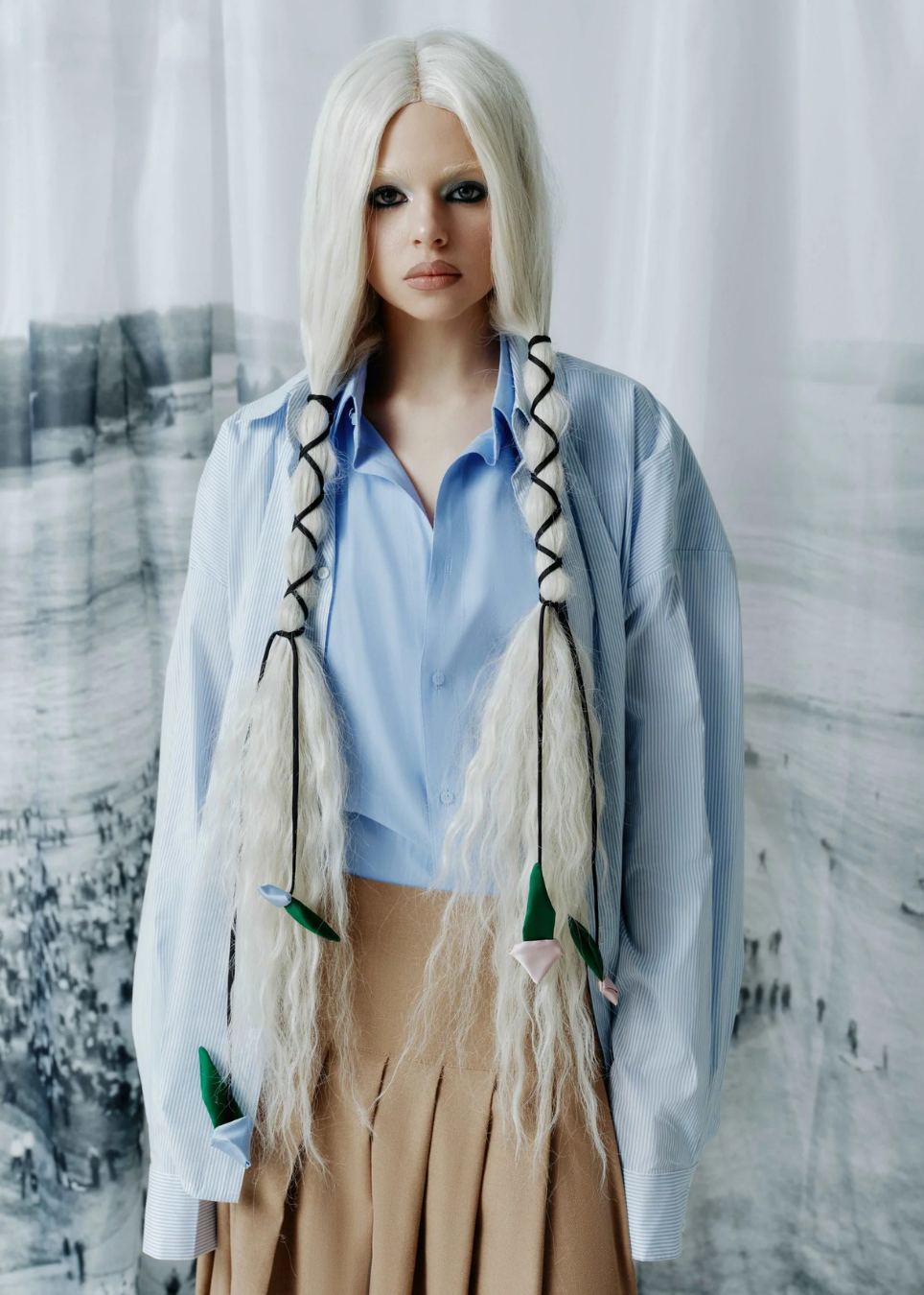 A model with long platinum blonde hair styled in two braids, wearing a light blue button-down shirt and a beige pleated skirt, posed against a white curtain backdrop.