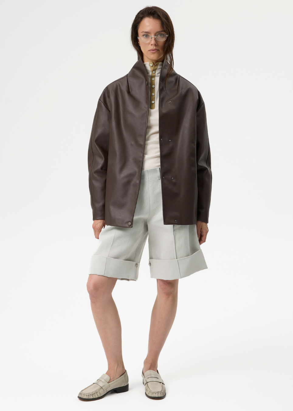 A model stands against a plain white studio background wearing a dark brown leather jacket over a cream shirt, light gray shorts, and beige loafers.