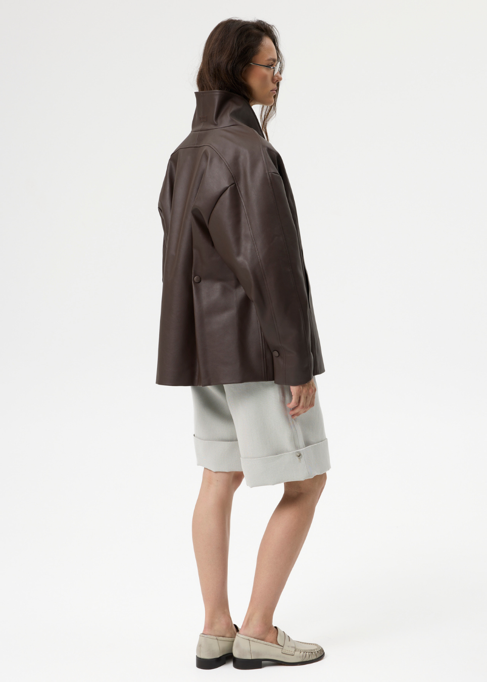 A model is shown from a side-back angle wearing a dark brown leather jacket, light gray rolled shorts, and beige loafers against a plain white studio background.
