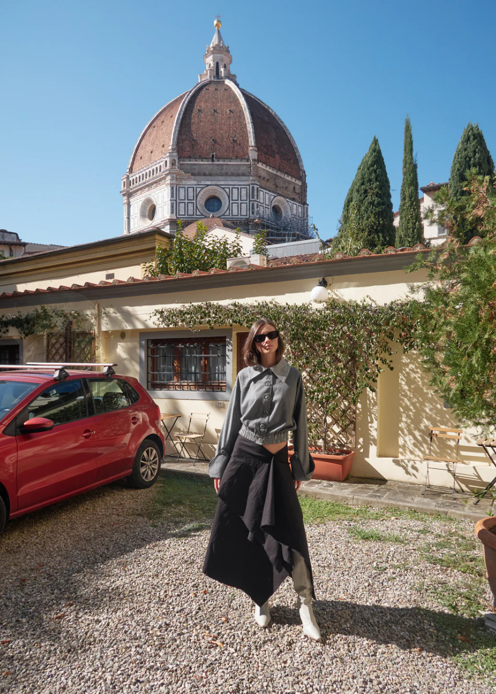 A woman wearing a gray long-sleeve top and a black asymmetrical skirt stands on a gravel path in front of a building with a large terracotta dome in the background, with a red car partially visible on the left.