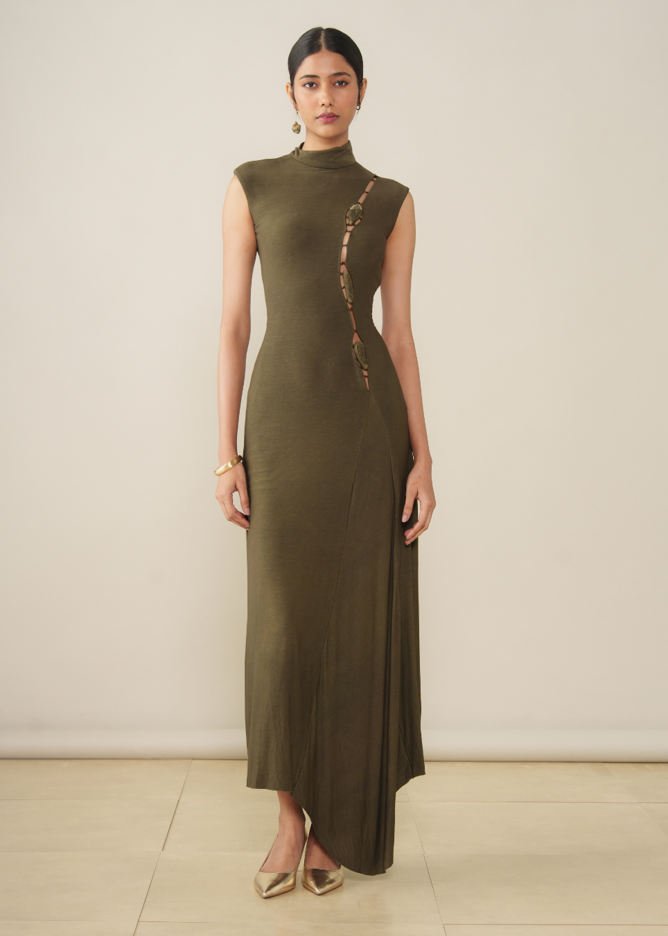 A woman modeling a sleeveless, high-neck, floor-length olive green dress with a vertical embroidered detail on the side, standing against a plain beige wall.