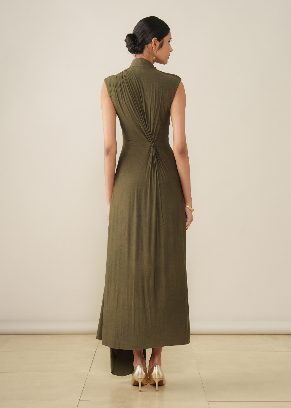 A woman stands with her back to the camera wearing a sleeveless, high-neck, floor-length olive green dress with dramatic gathered pleating at the back waist. She is wearing gold heels against a plain beige studio background.