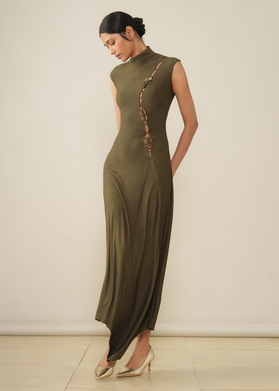 A woman modeling a sleeveless, high-neck, floor-length olive green dress with a decorative metallic pattern running down the side, paired with beige pointed-toe heels against a plain cream background.