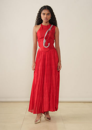 A model wearing a vibrant red sleeveless maxi dress with a pleated skirt and a grey embellished bodice, standing against a plain beige studio background.
