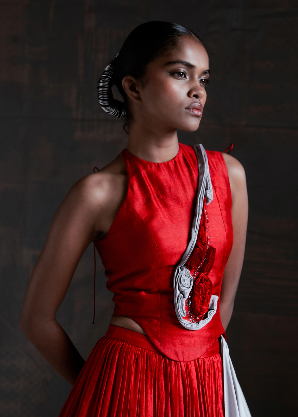 A model wearing a vibrant red sleeveless top and matching pleated skirt, styled with a white and red patterned shoulder bag, set against a dark textured studio background.