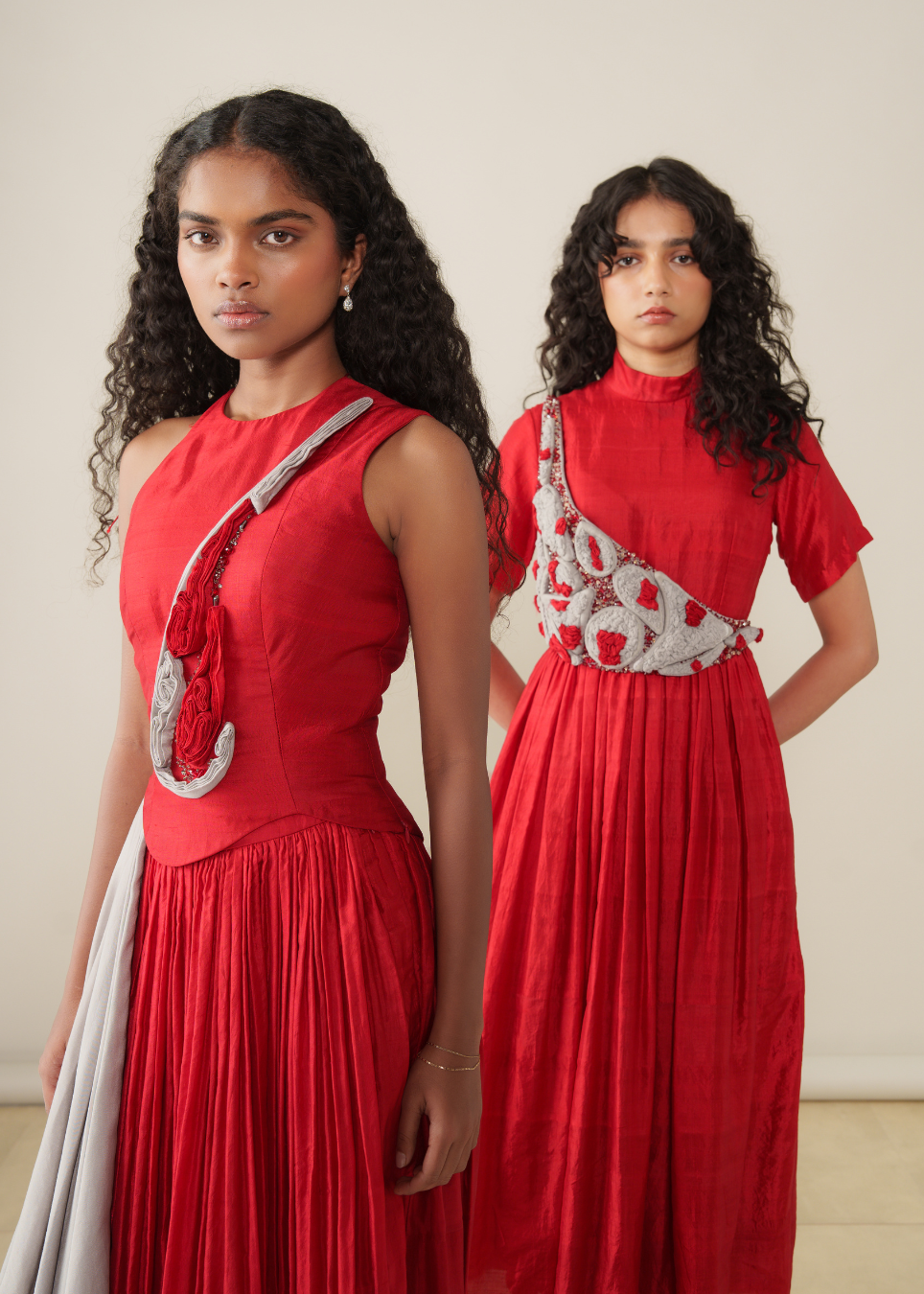 Two models wearing red sleeveless maxi dresses with a draped silver-gray fabric panel across the chest, posed against a plain beige studio background.