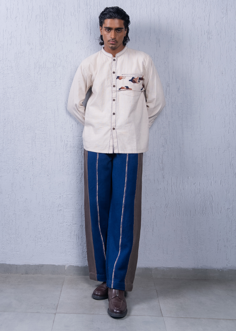 A man modeling a cream-colored long-sleeve kurta with a floral chest pocket detail, paired with blue trousers featuring brown side panels and white stripes, standing against a textured light gray wall.