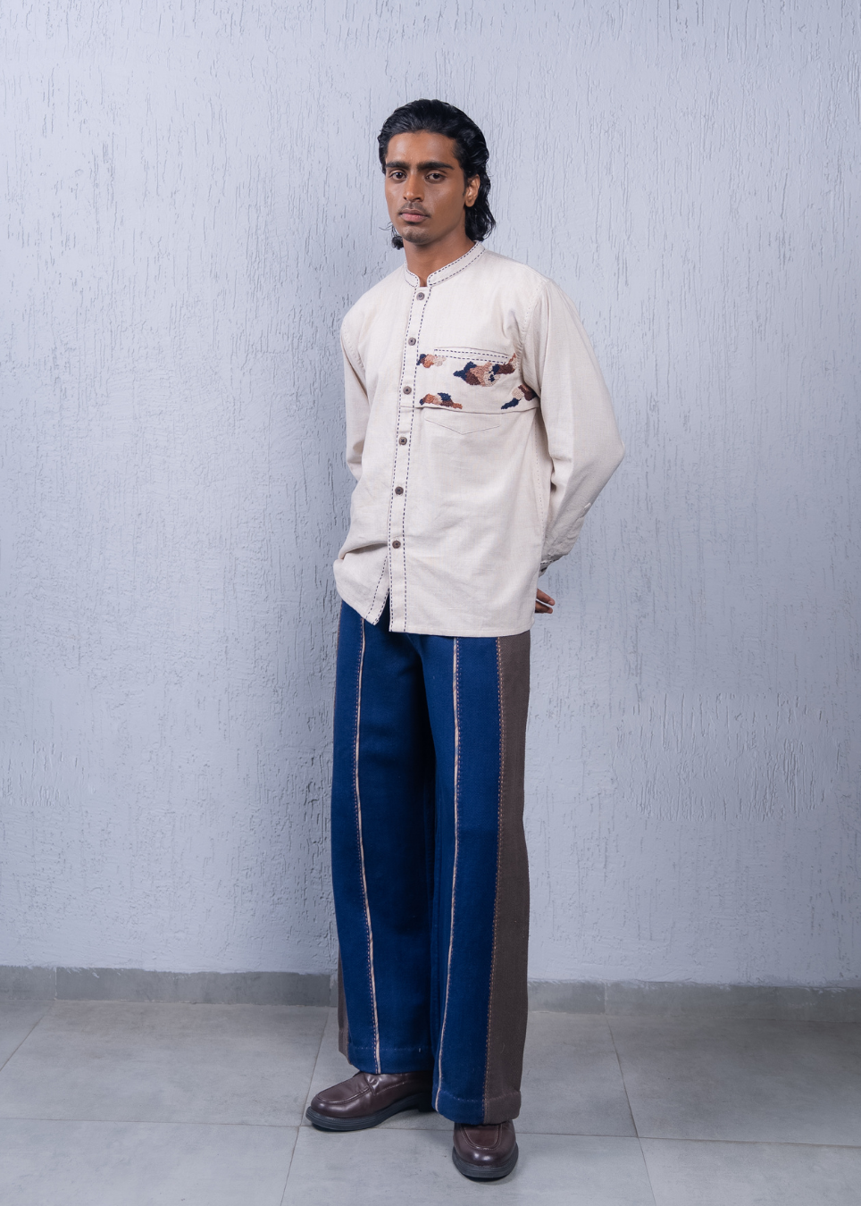 A man modeling a cream-colored long-sleeve button-up shirt with a colorful bird pattern on the pocket, paired with blue and brown side-striped trousers and brown leather shoes, standing against a textured light gray wall.