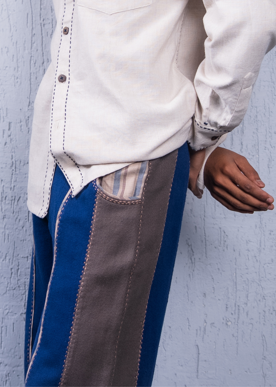 A close-up of a person wearing a cream-colored shirt tucked into blue and taupe patchwork trousers, standing against a textured light blue-gray wall.