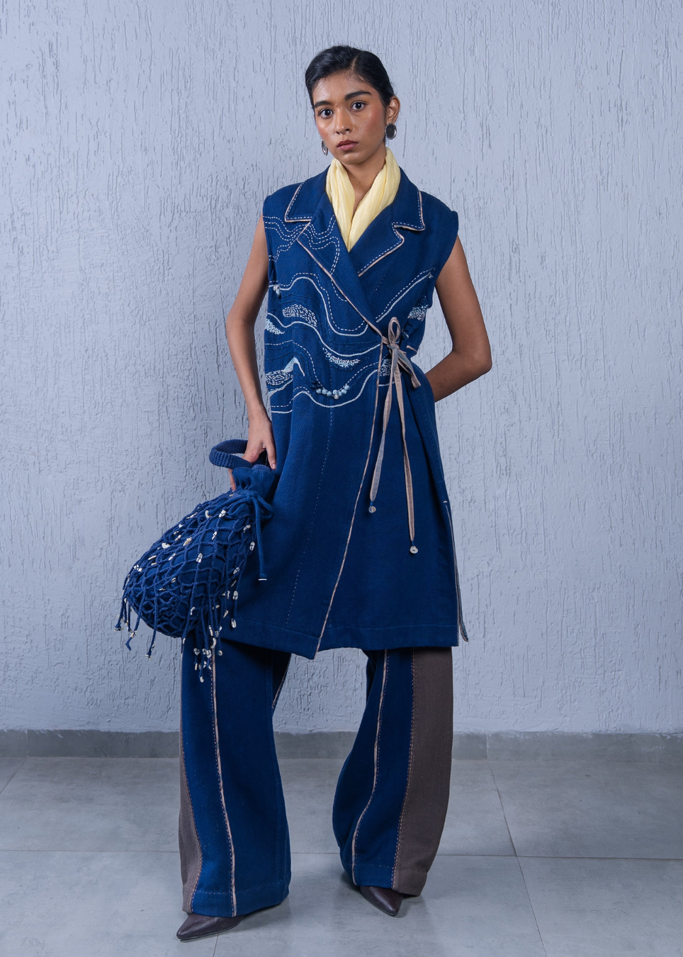 A model stands against a textured light gray wall wearing a blue sleeveless wrap-style tunic with white wave embroidery, matching wide-leg pants with blue and brown side panels, a yellow scarf, and holding a blue beaded fringe bag.