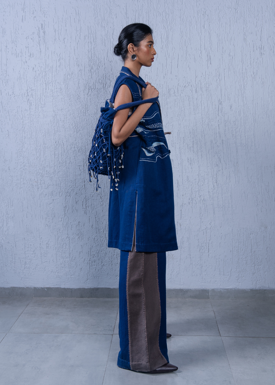 A model is shown from the side profile wearing a dark blue indigo tunic with white embroidery and matching wide-leg trousers with brown side panels, holding a blue patterned shawl against a textured light gray wall.
