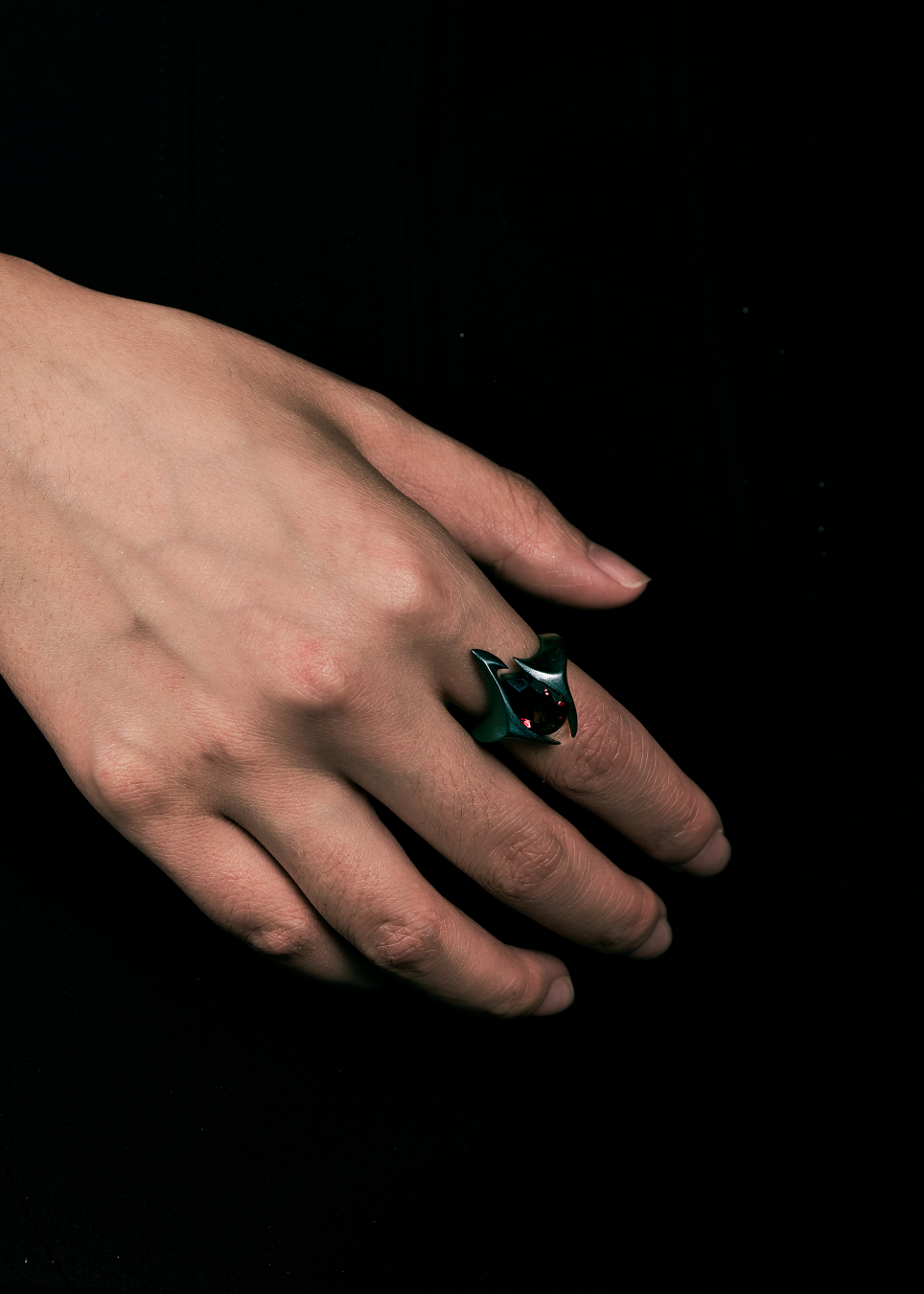 A close-up of a hand wearing a sculptural dark green ring with a black and white bird-like face, set against a solid black background.