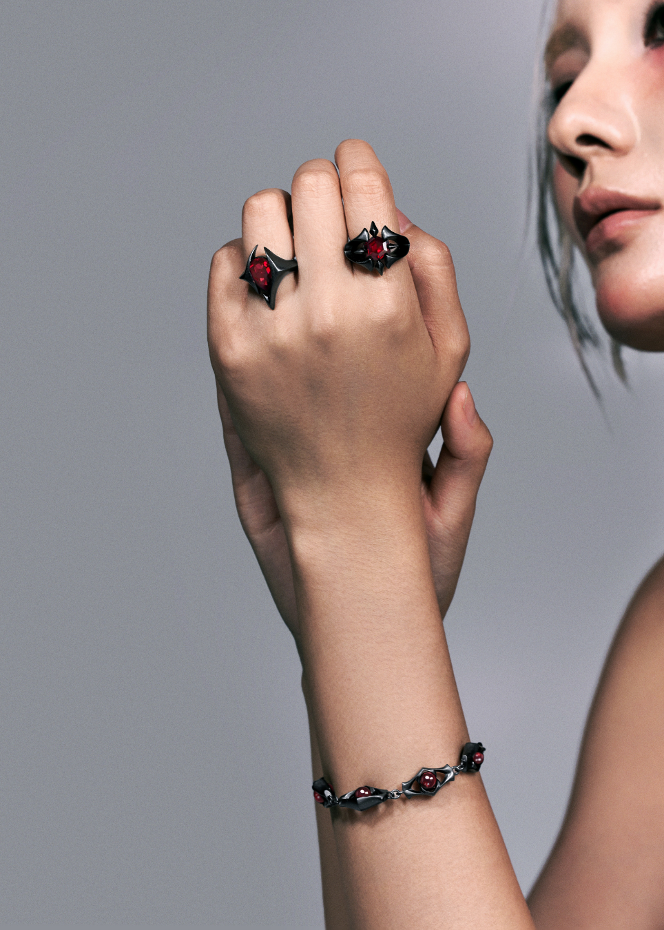 A close-up of a model's hand and wrist wearing two matching gothic-style rings and a matching bracelet. The jewelry features dark, starburst-shaped settings with deep red gemstones, set against a plain gray studio background.