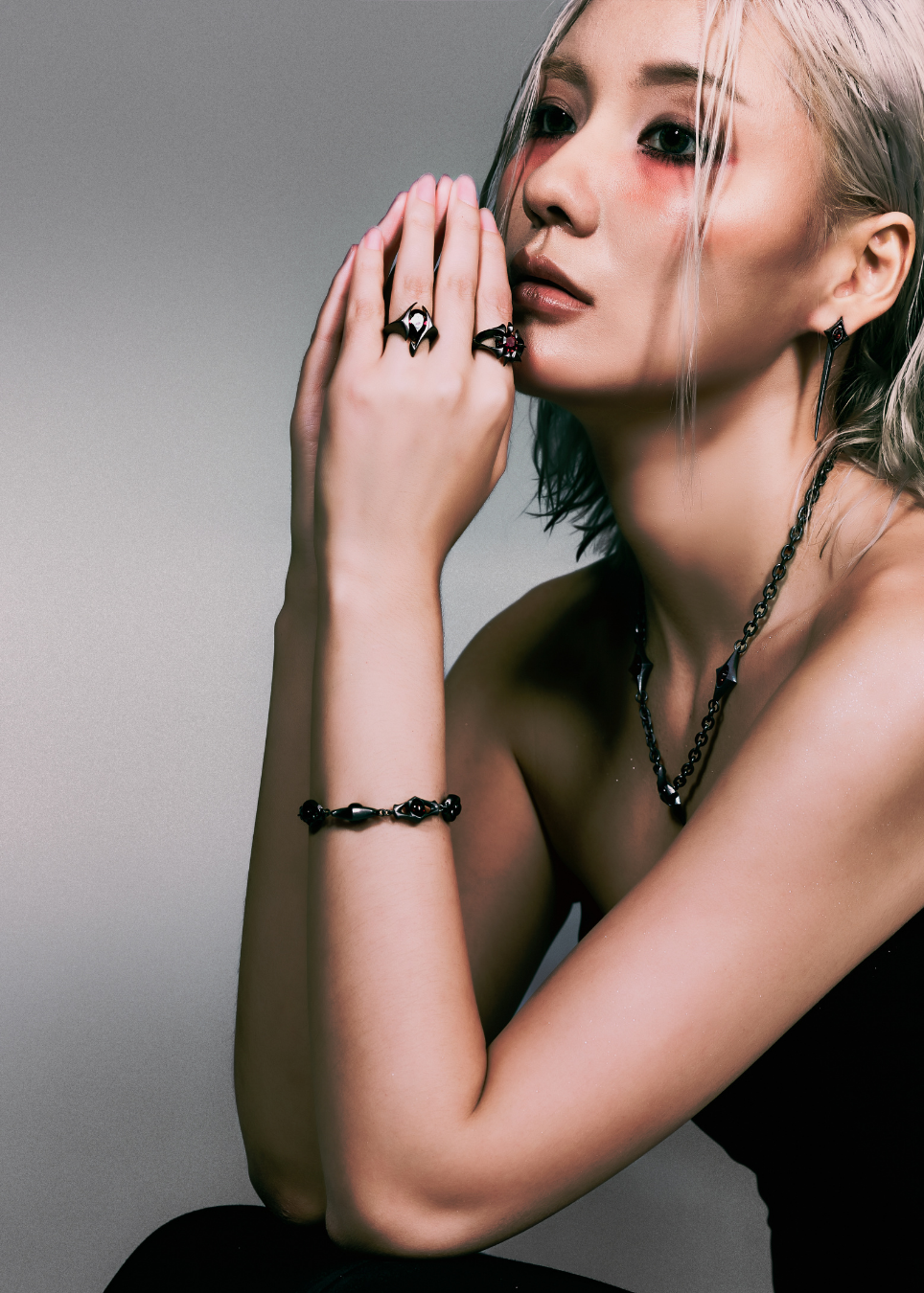 A close-up portrait of a woman with platinum blonde hair wearing black jewelry, including a black beaded bracelet, a black chain necklace, and black rings on her fingers, posed against a neutral gray background.